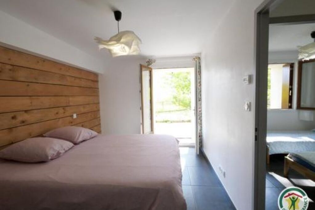 A bedroom with a wooden headboard, a bed with a light-colored bedspread, and a window with a view of greenery outside.