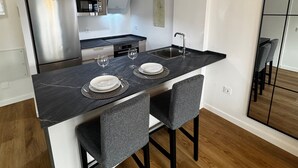 A modern kitchen with a black countertop, two grey upholstered chairs, and a stainless steel refrigerator.