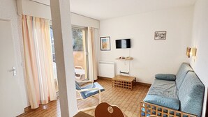 Ein gemütlicher, kompakter Wohnraum mit Holzboden, einer Couch, einem Esstisch, einem an der Wand befestigten Fernseher und einem Fenster mit Vorhängen.