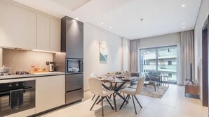 A modern kitchen with a built-in oven and microwave, a dining area with a glass table and chairs, and a view of a high-rise building through large windows. - Manzil - 1BR in Arjan with City Views (Dubai)