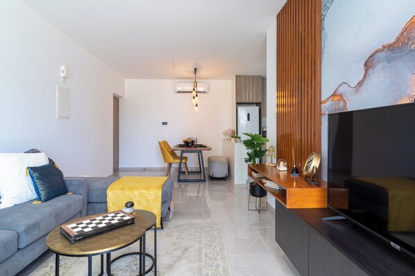 A modern living room with a sofa, coffee table, and a TV. There is a dining area with a table and chairs. - Flair 1-BR Apt. & Balcony in Larnaca (Larnaca)