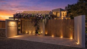 Een moderne villa met een goed verlichte entree, een houten hek en een bord met de tekst 'CASA HARMONIA'.