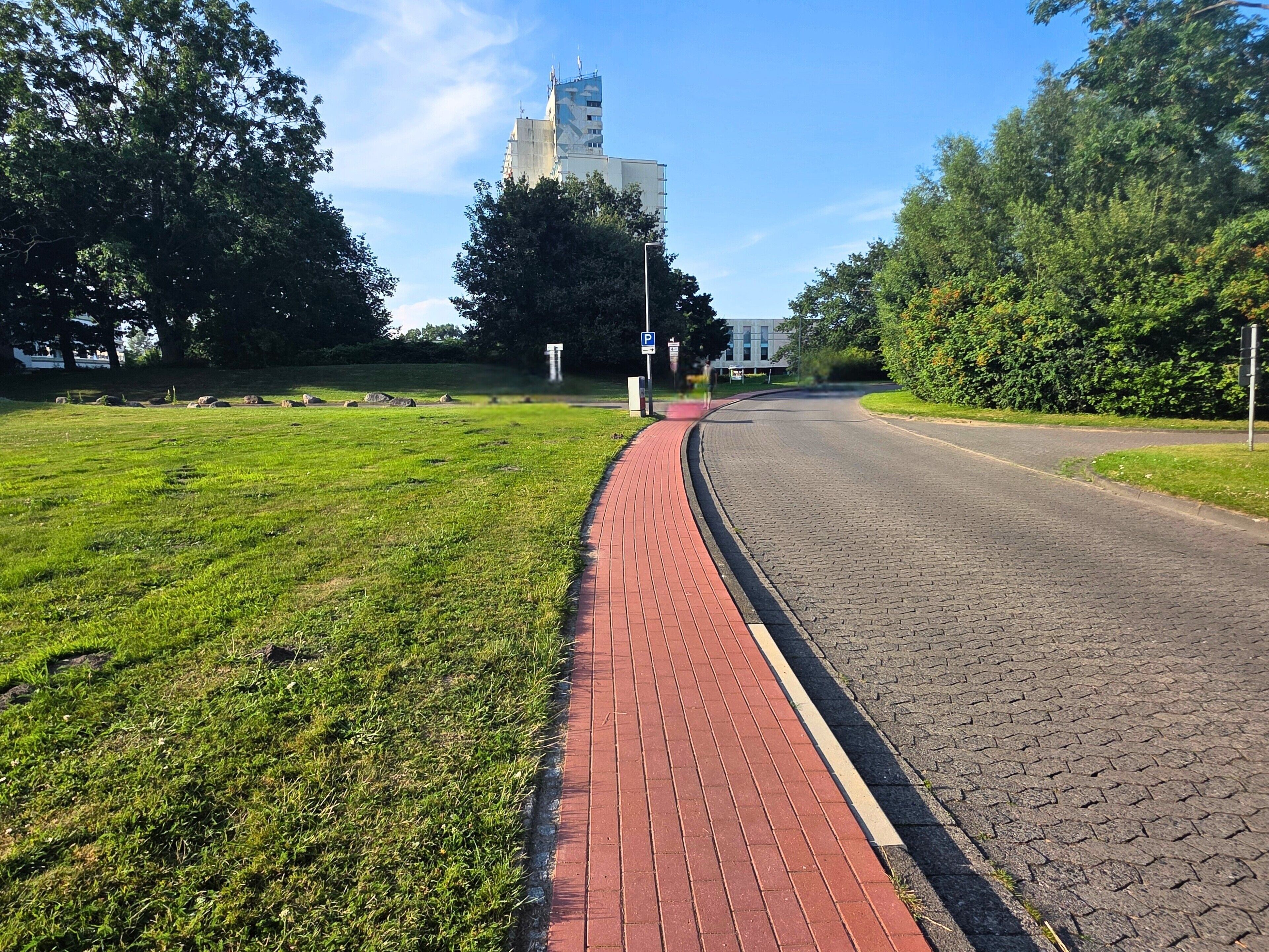 A paved road with a red brick sidewalk, green grass, trees, and a distant building.