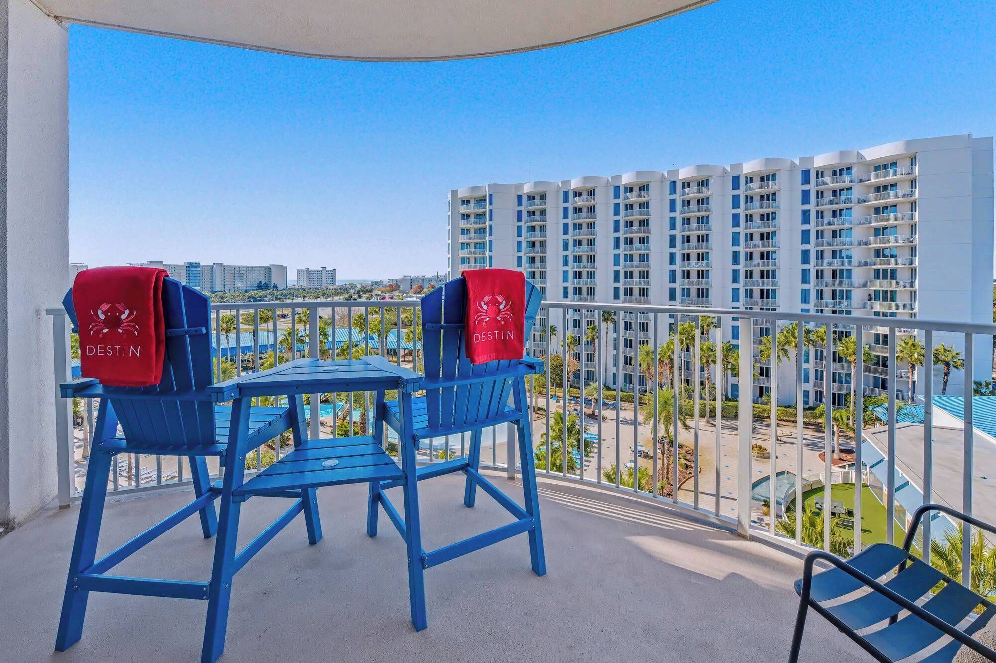 A balcony with two blue chairs and a table, overlooking a pool and a high-rise building.