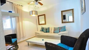 Una sala de estar con un sofá, dos sillones de mimbre, una mesa de centro, un ventilador de techo y un televisor fijado a la pared.