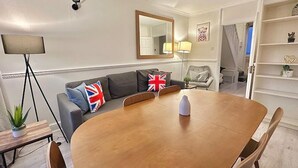 Un salon moderne avec une table à manger en bois, un canapé gris orné de drapeaux britanniques, un miroir et une bibliothèque.