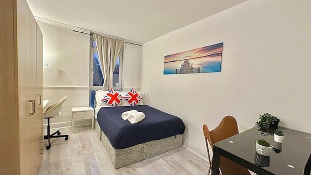 Une chambre d’hôtel avec un lit, un bureau, une chaise, une fenêtre avec des rideaux, un tableau au mur et une petite table avec une plante.