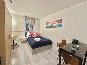 A hotel room with a bed, a desk, a chair, a window with curtains, a painting on the wall, and a small table with a plant. - Cosy Abbey Road Apartment (London)