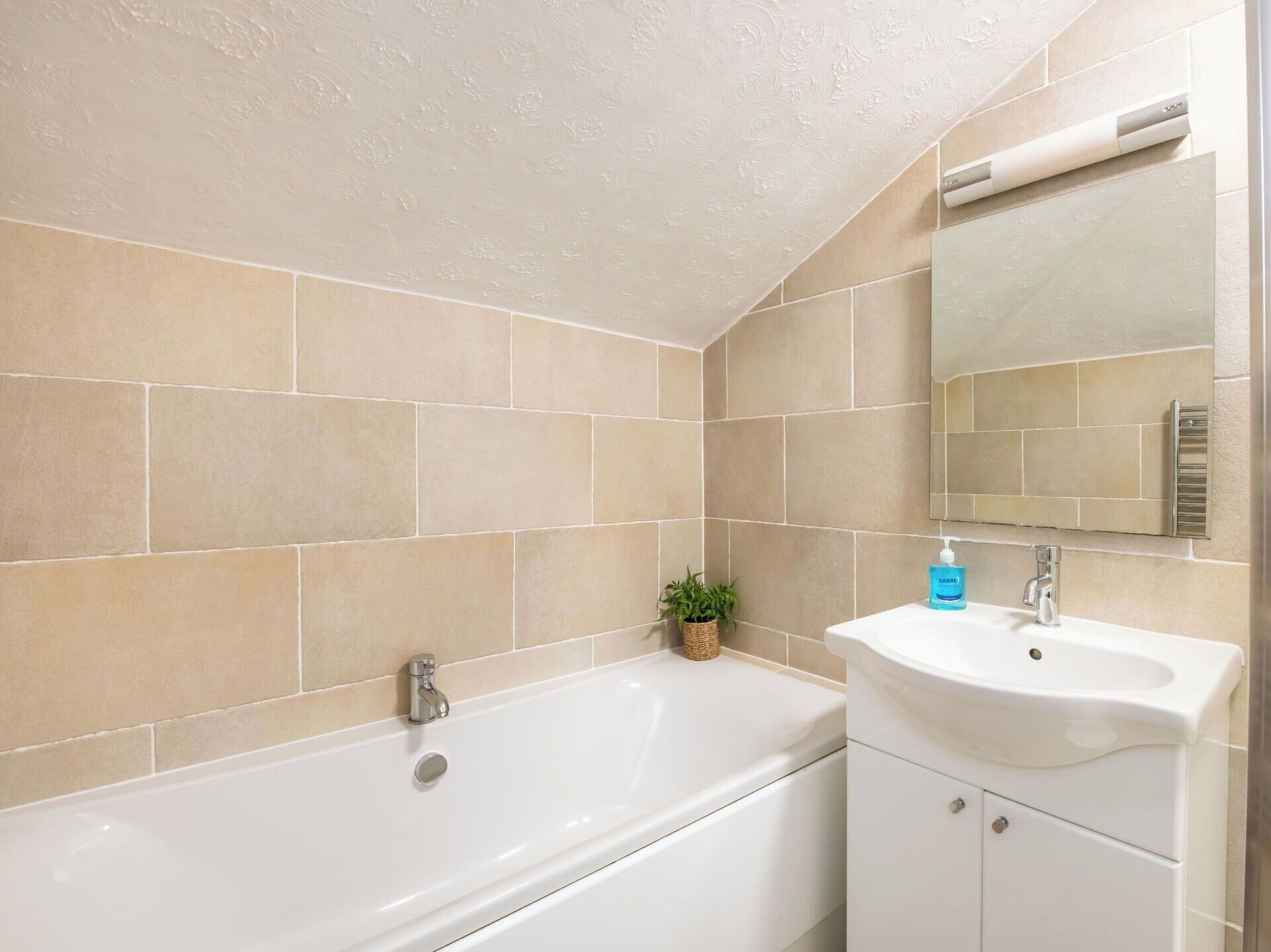 Un baño con baldosas color beige, una bañera blanca, un lavamanos blanco y una pequeña planta en maceta.