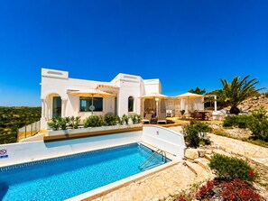 A modern white villa with a swimming pool, outdoor seating, and a view of the landscape.