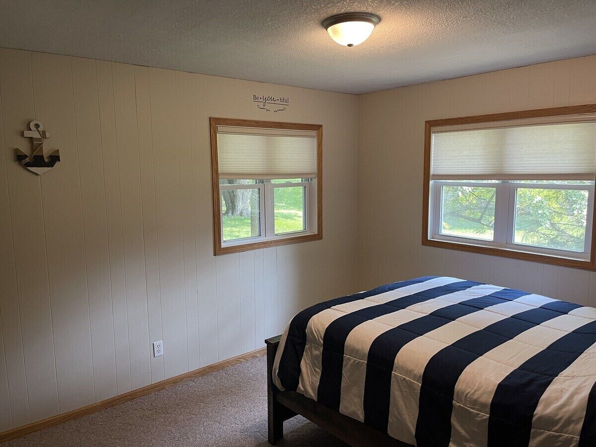Ein Schlafzimmer mit zwei Fenstern, einem Bett mit gestreifter Bettdecke und einer wandmontierten Lampe.