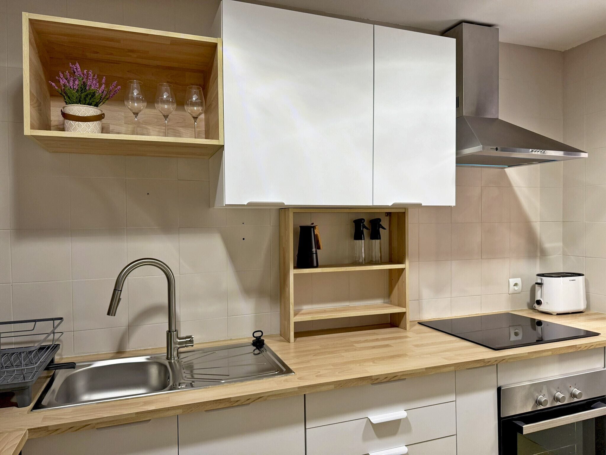 Una cocina moderna con un lavamanos de acero inoxidable, encimera de madera, armarios blancos, horno empotrado y campana extractora.