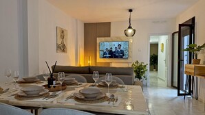 Un comedor moderno con una mesa larga dispuesta para una comida, un televisor montado en la pared y un sofá al fondo.