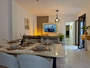 Un comedor moderno con una mesa larga dispuesta para una comida, un televisor montado en la pared y un sofá al fondo.