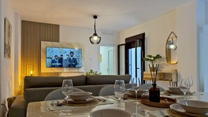 Un comedor con una mesa dispuesta para cuatro, un televisor montado en la pared y un sofá al fondo.