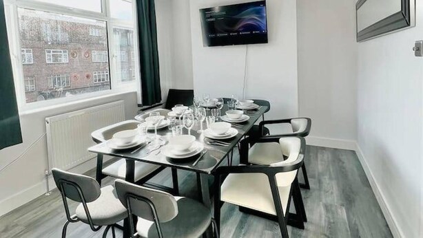 A modern dining room with a long table set for six, equipped with chairs, plates, glasses, and cutlery. A flat-screen TV is mounted on the wall above the table. - Sunny Neasden Apartment (London)