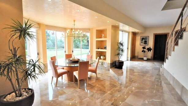 A spacious dining area with a round table and chairs, a chandelier, and potted plants. - Sa Rota | Pool, Tennis & Nature (BUNYOLA)