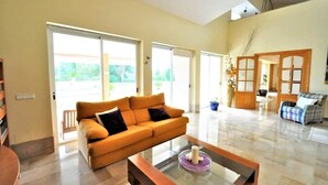 A spacious living room with a large sofa, a coffee table, and a view of the outdoors through large windows. - Sa Rota | Pool, Tennis & Nature (BUNYOLA)