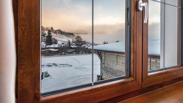 Blick durch ein Fenster mit Holzrahmen auf eine verschneite Landschaft mit Häusern und Bäumen.