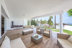 A modern outdoor living area with wicker furniture, a glass coffee table, and a view of palm trees and buildings. - Villa "Jazz Close To Golden Mile" with Sea View, Private Pool & Wi-Fi (Marbella)
