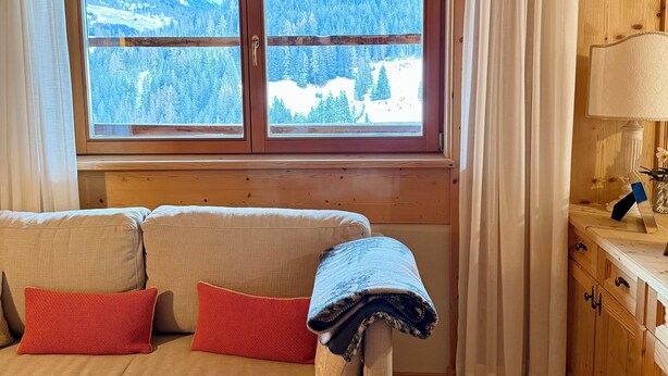 A cozy room with a wooden ceiling, a sofa with cushions, a window with a view of a snowy landscape, and a lamp on a wooden table.