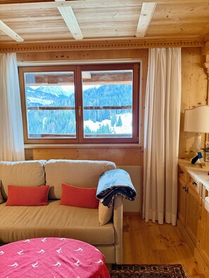 Ein gemütliches Zimmer mit Holzbalkendecke, einer Couch mit Kissen, einem Fenster mit Blick auf eine verschneite Landschaft und einer Lampe auf einem Holztisch.