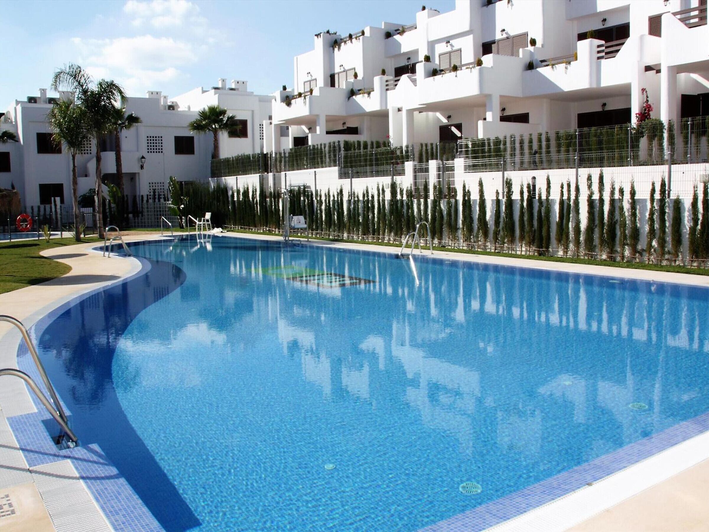 Une piscine à l’eau d’un bleu cristallin, entourée d’un bâtiment blanc doté de balcons et bordée d’une rangée d’arbres.