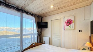 A bedroom with a large window, a bed, a flat-screen TV, and a painting of a flower. - Renovated 2-room apartment - 7 ppl - Ski-in/Ski-out (Le Corbier)