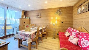 A cozy room with a wooden interior, a dining table with chairs, a red sofa with patterned cushions, and a wall-mounted lamp. - Renovated 2-room apartment - 7 ppl - Ski-in/Ski-out (Le Corbier)