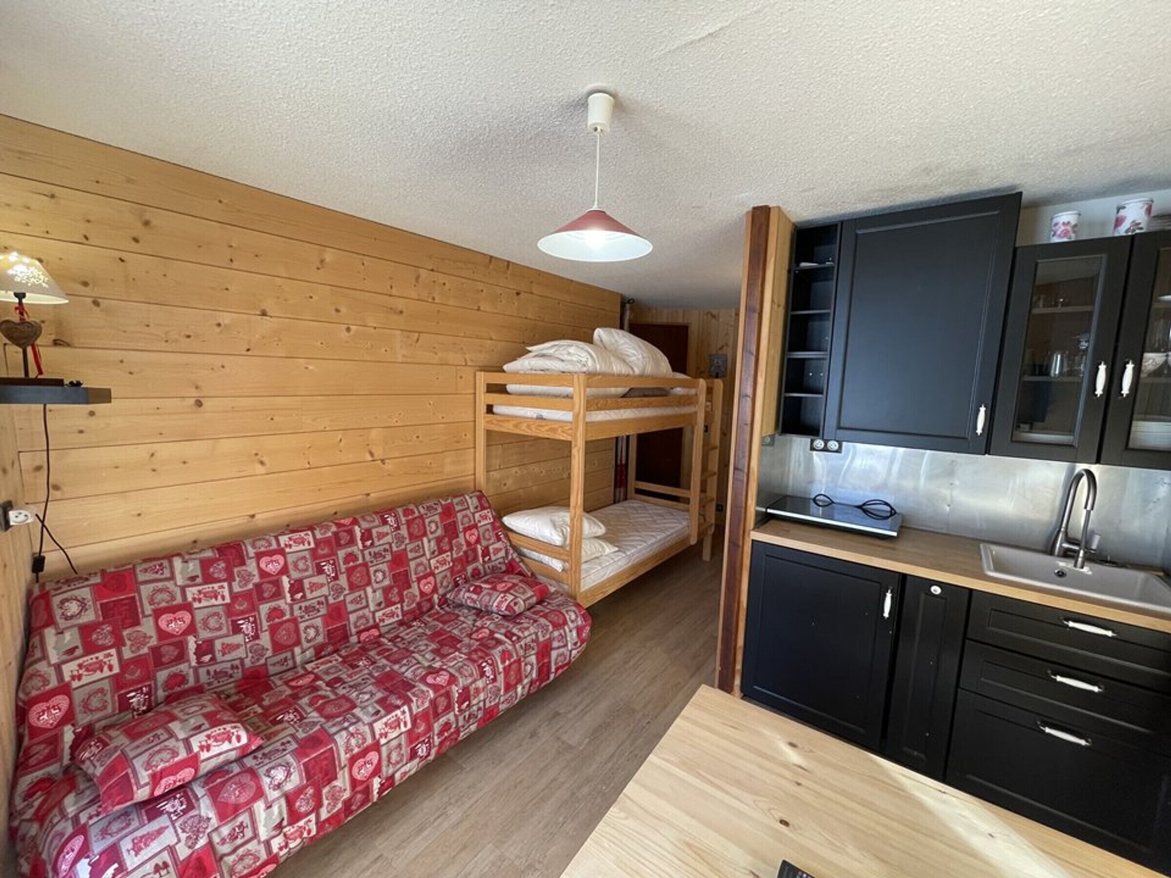Et lille værelse med køjeseng, sofa med rødt mønster, lille tekøkken med vask og trævægge.