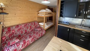 A compact room with a bunk bed, a sofa with a red patterned cover, a small kitchenette with a sink, and wooden walls. - Charming renovated studio with balcony in Lioran (Le Lioran)