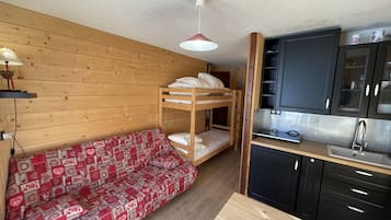 Et lille værelse med køjeseng, sofa med rødt mønster, lille tekøkken med vask og trævægge.