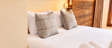 A neatly made bed with white linens and two patterned pillows, a framed picture above it, and a rolled towel on the side.