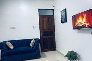 A living room with a blue sofa, a wooden door, a TV displaying a fire, and a potted plant. - Sea Side Residence – Your Cozy Coastal Retreat with Stunning Views (Mombasa)