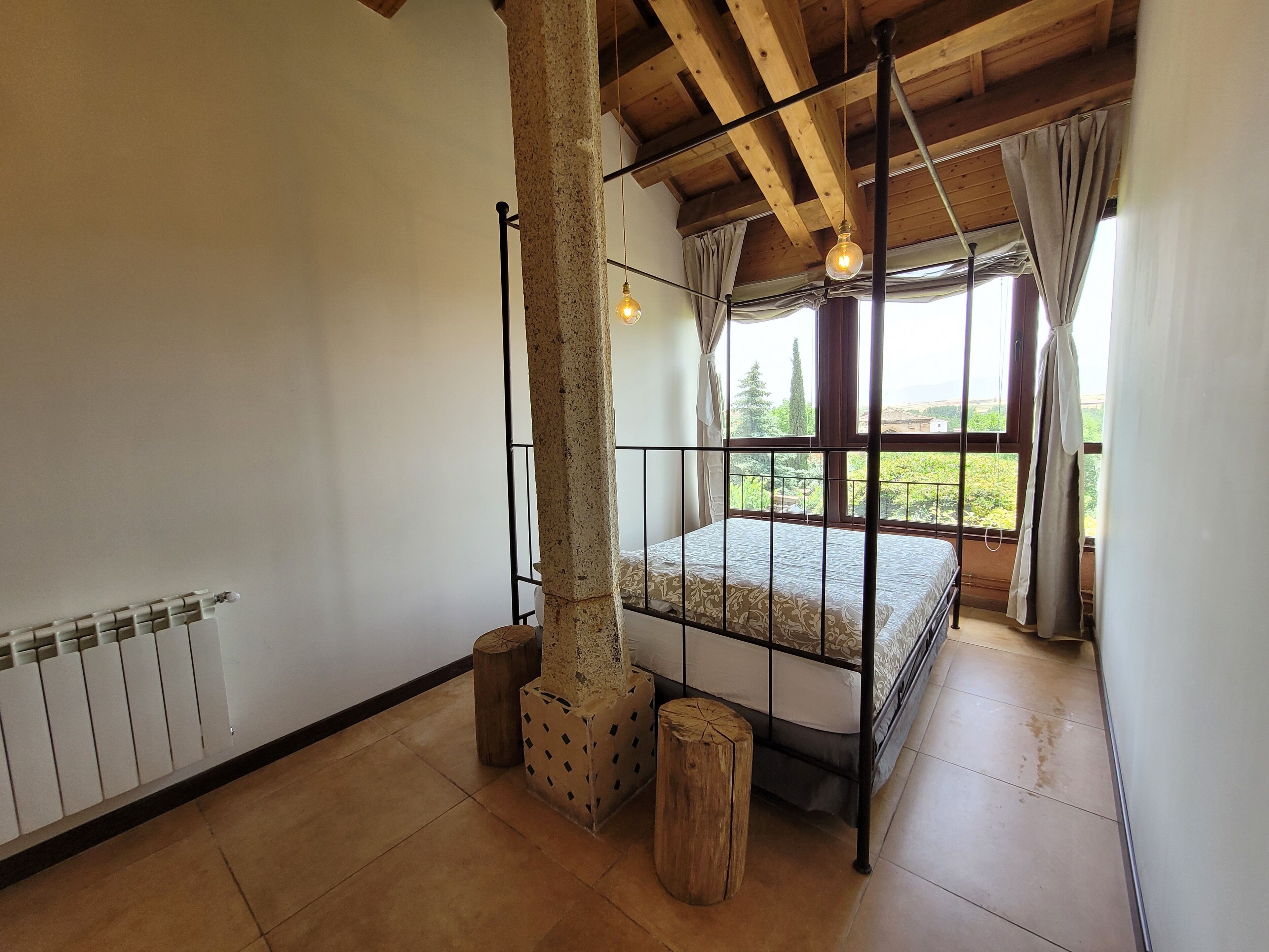 A bedroom with a wooden ceiling, a bed with a metal frame, a radiator, and a view of greenery outside.