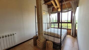 A bedroom with a wooden ceiling, a bed with a metal frame, a radiator, and a view of greenery outside.