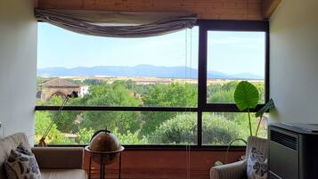 A room with a large window offering a view of a landscape with mountains and greenery. There is a sofa, a chair, and a table in the room.