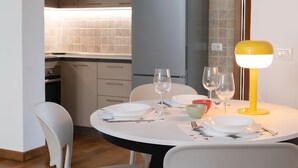 A dining area with a round table set for two, two white chairs, a yellow table lamp, and a grey refrigerator in the background. - Sea view holiday home, surrounded by Mediterranean scrub with swimming pool (Chioma)