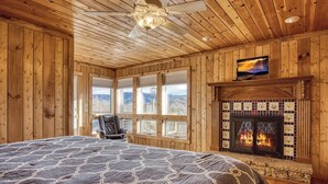 Een slaapkamer in een houten chalet met een groot bed, een open haard, een televisie en uitzicht op de bergen door de ramen.