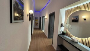 Ein Flur mit gerahmten Kunstwerken, Wandleuchten, Holzboden und einem geschwungenen Spiegel mit Holzpaneelen.