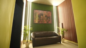 Une pièce avec un mur vert, un canapé en cuir marron et un grand tableau représentant des motifs floraux. Deux plantes en pot sont placées de part et d’autre du canapé.