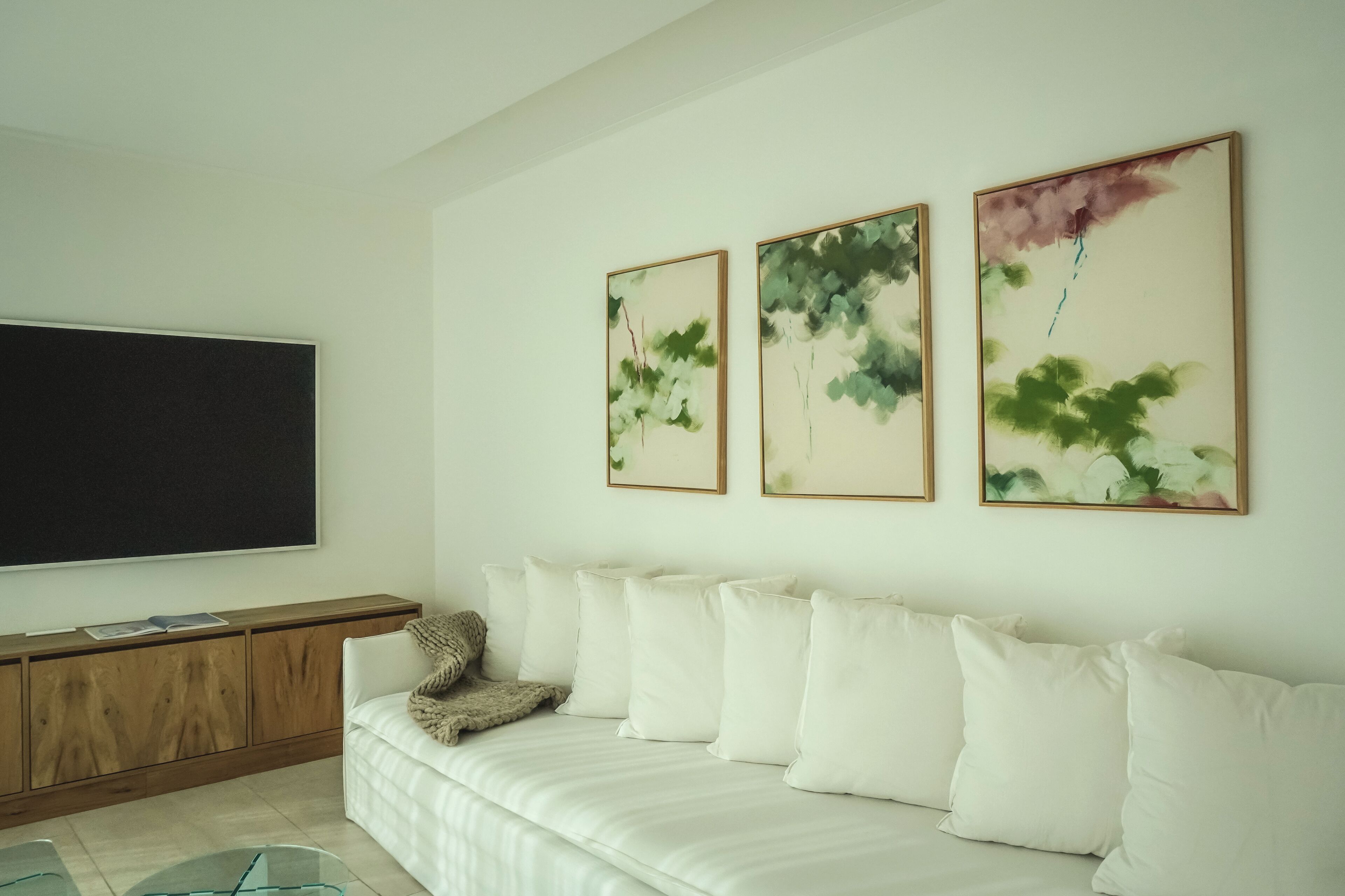 A modern living room with a flat-screen TV, a wooden cabinet, and a white sofa with multiple cushions.