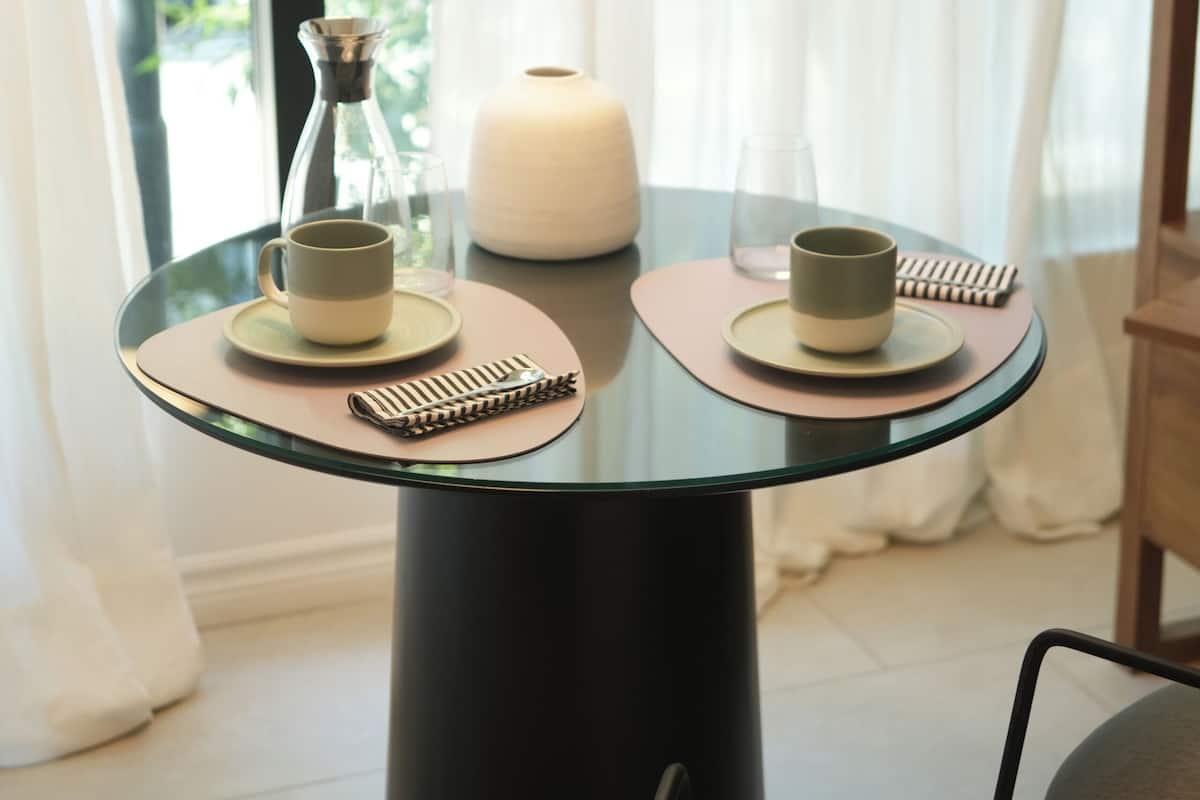 Une table en verre avec deux tasses et une bougie, placée devant une fenêtre ornée de rideaux.