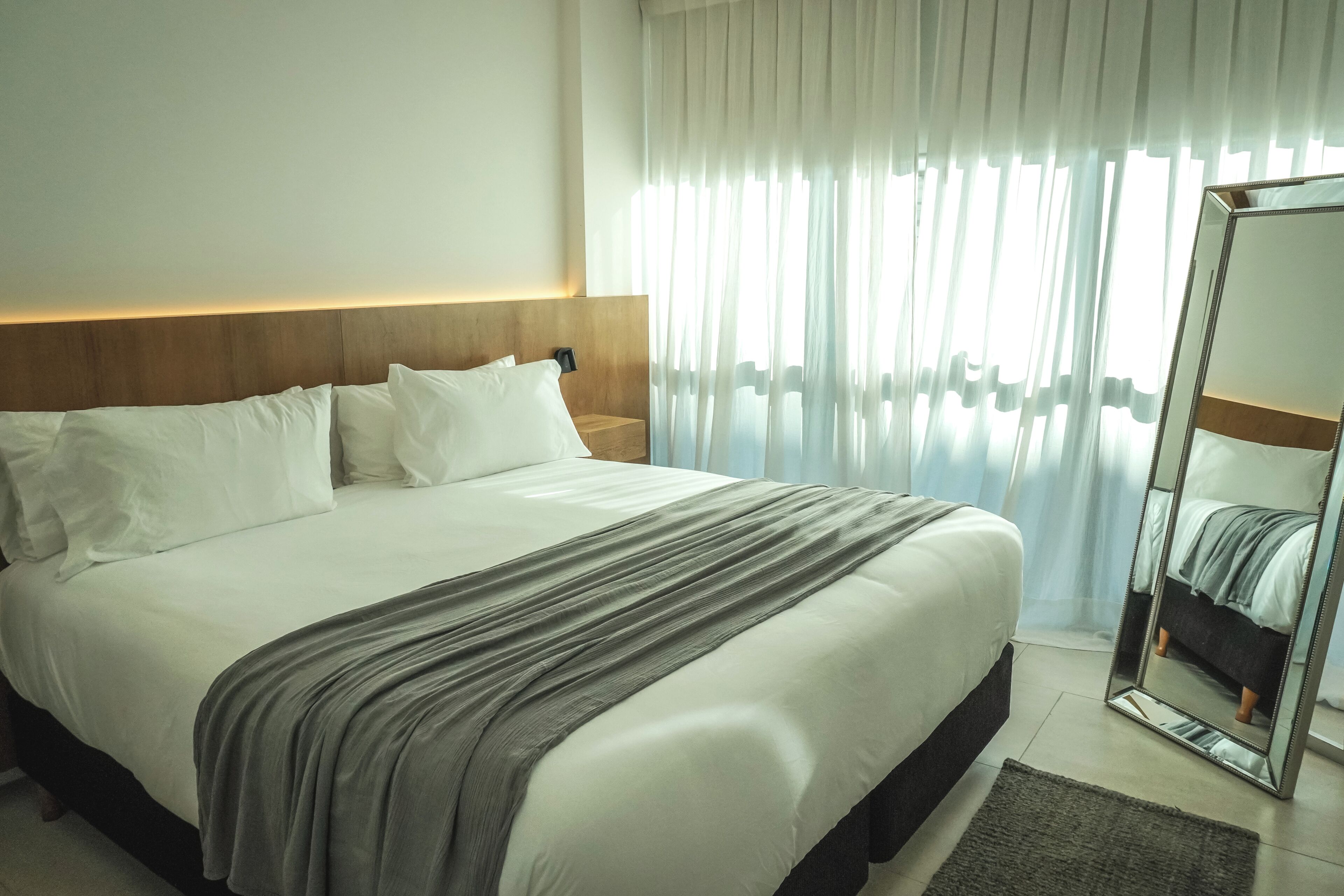 Habitación de hotel con una cama grande, cabecero, mesita de noche, espejo y cortinas traslúcidas.