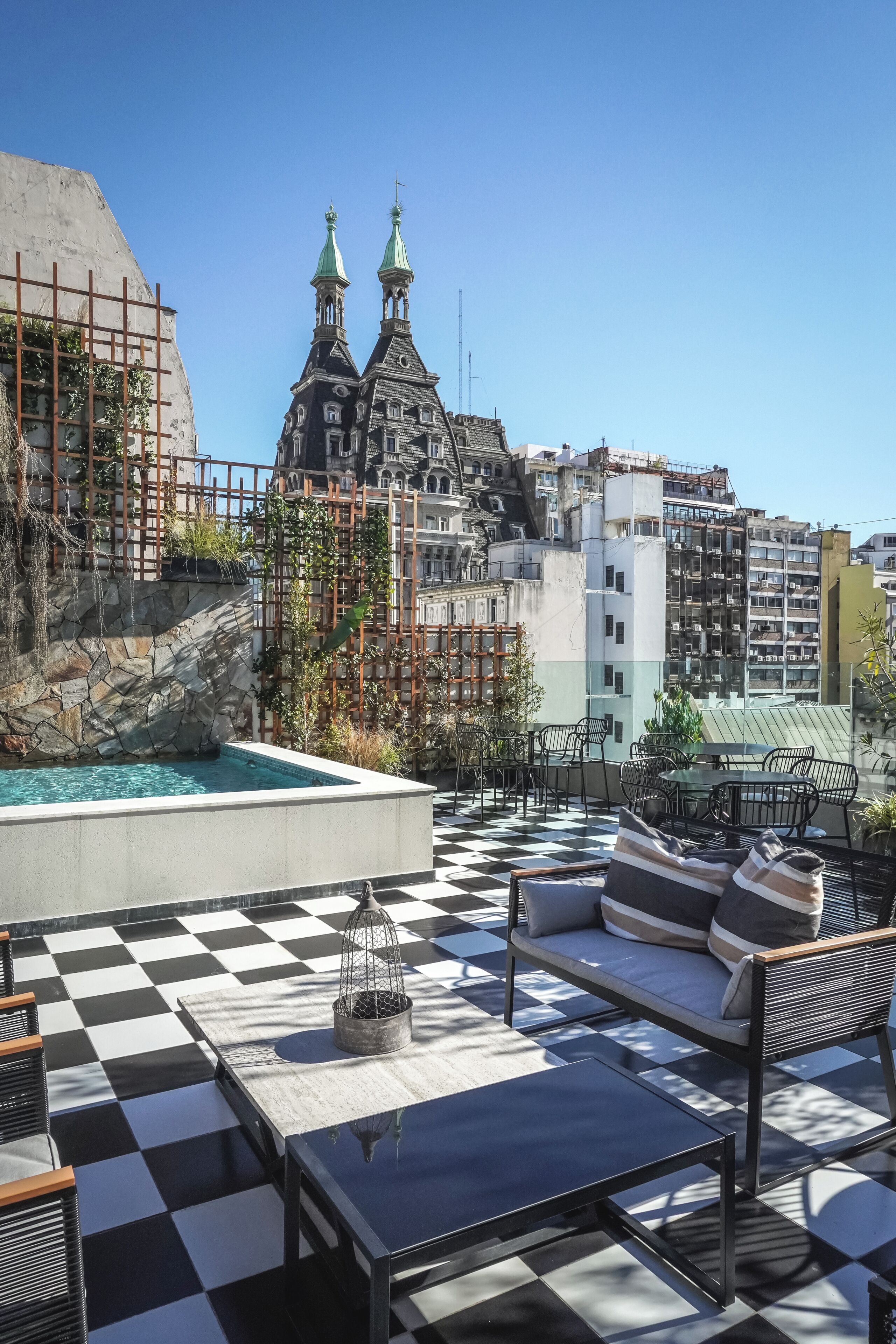 A rooftop terrace with a checkered floor, outdoor seating, a pool, and a view of historic buildings with spires.