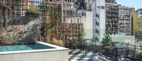 Une terrasse sur le toit avec un sol à carreaux, des sièges en plein air, une piscine et une vue sur des bâtiments historiques dotés de flèches.