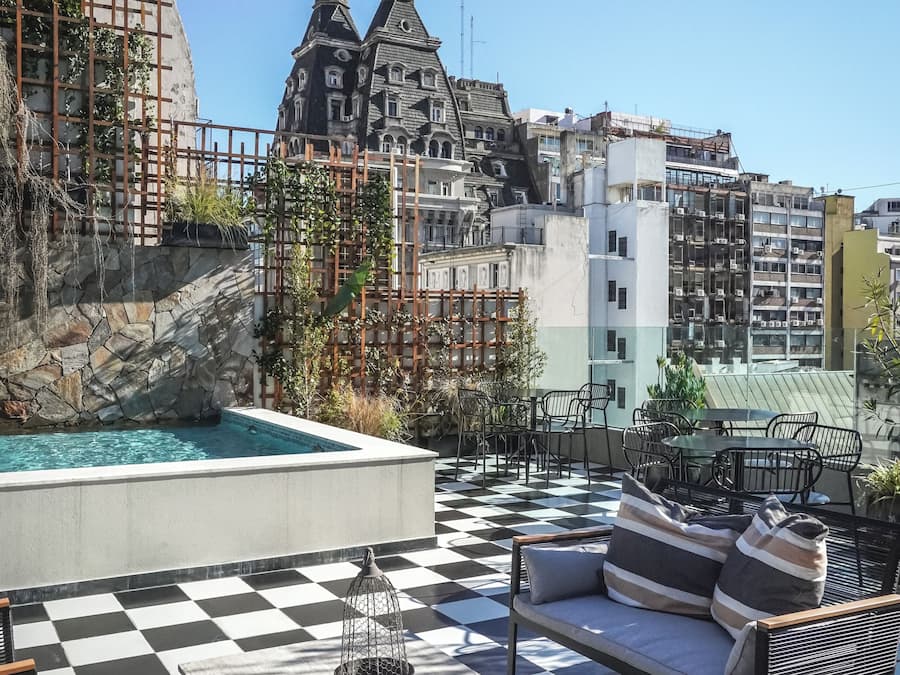 Une terrasse sur le toit avec un sol à carreaux, des sièges en plein air, une piscine et une vue sur des bâtiments historiques dotés de flèches.
