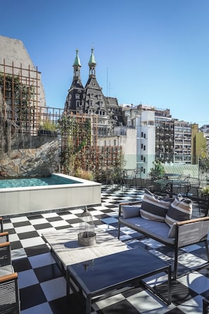 Une terrasse sur le toit avec un sol à carreaux, des sièges en plein air, une piscine et une vue sur des bâtiments historiques dotés de flèches.
