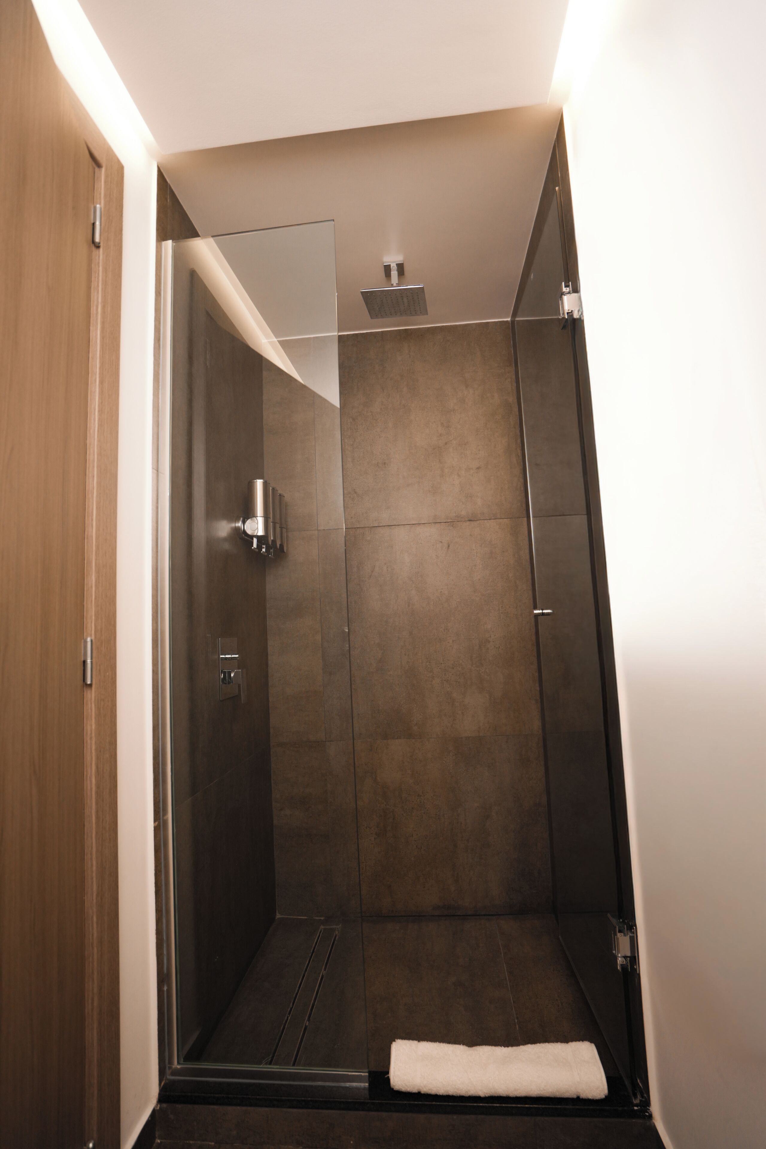 A modern shower with a glass door, a towel on the floor, and a wooden door to the left.