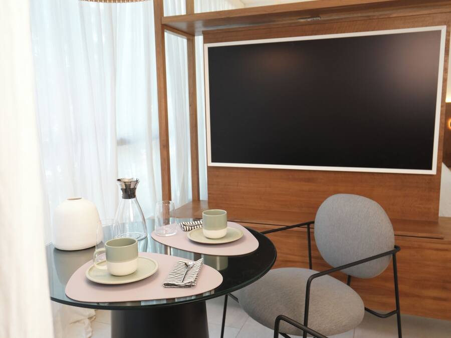 Un comedor moderno con una mesa redonda de vidrio, dos sillas grises tapizadas, una lámpara colgante negra y un televisor de pantalla plana grande montado en la pared.